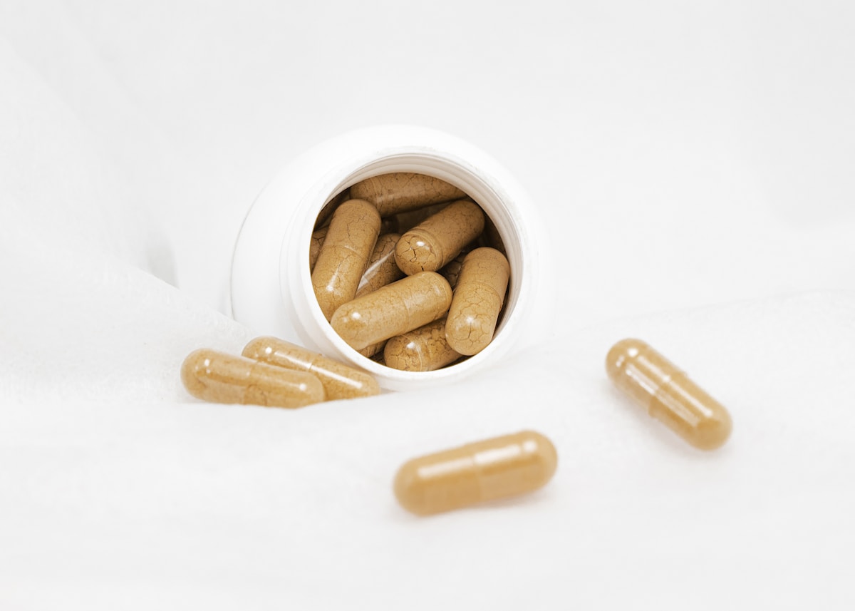 Natural supplement capsules spilling from a white bottle with green herbs — clean label lubricant